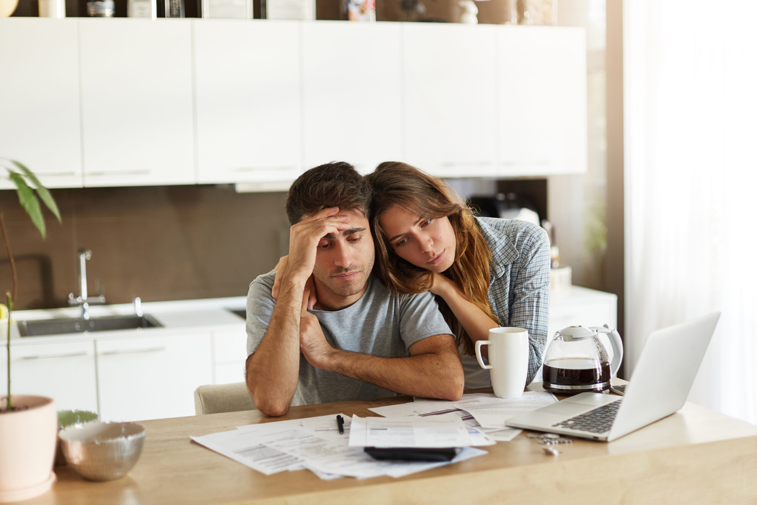 Stressed wife and husband with many debts doing paperwork together, reviewing their expenses, planning domestic budget and calculating bills in kitchen, surrounded with papers, calculator and laptop